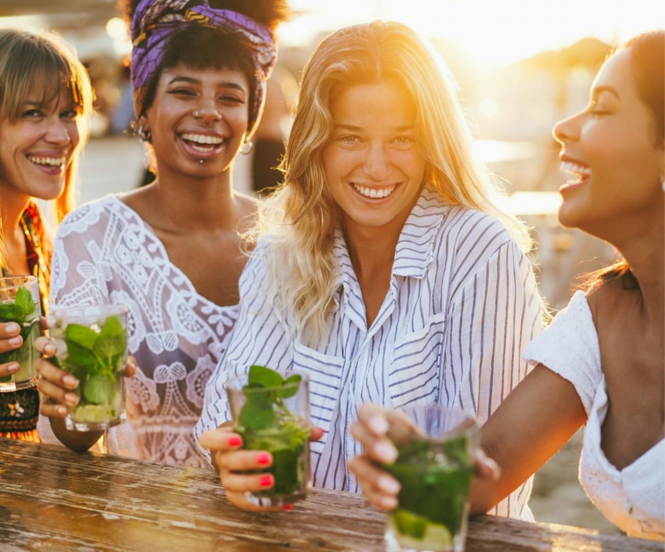 Quattro amiche sorridenti bevono cocktail insieme al tramonto.