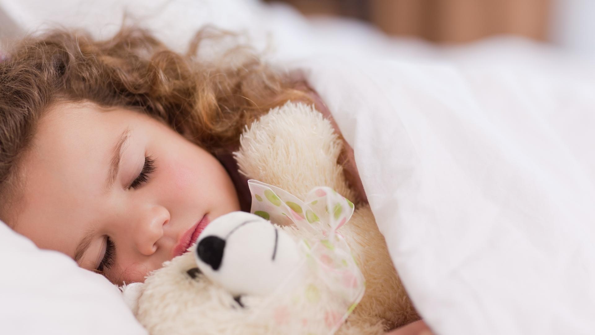 Child sleeping, hugging a stuffed toy under a white blanket.