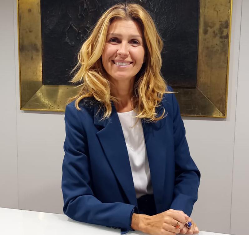 Donna sorridente in giacca blu davanti a un quadro scuro.